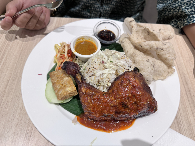 A plate of Nasi ulam with rice, grilled chicken, tofu, vegetables, and crackers, served with various sauces, with a person's hand visible holding a spoon.
