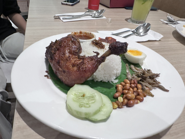 A plate of nasi lemak with rice, grilled chicken, cucumber slices, a boiled egg, peanuts, and small fried fish, served on a banana leaf. There are utensils and another diner in the background.