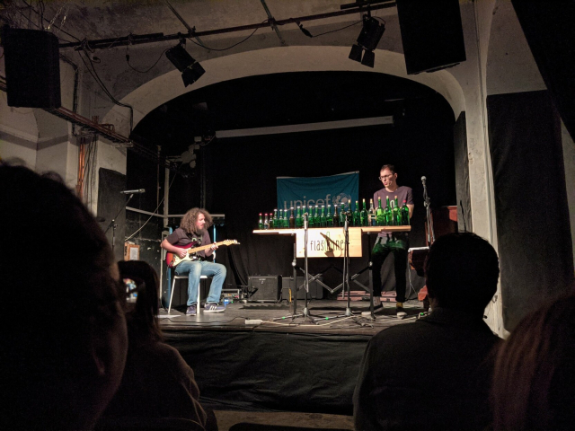 Flaškinet music band playing on the stage: one guy playing electric guitar and and second guy playing on a row of glass bottles.