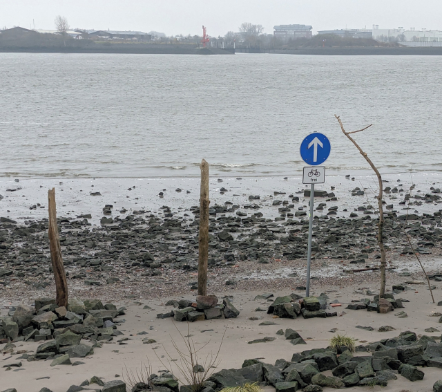 Im Elbstrand steckt ein Schild "geradeaus fahren, Fahrräder frei", das direkt auf die Elbe zeigt.