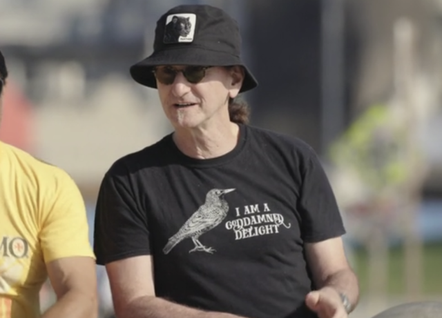 Geddy Lee standing in the sunlight and wearing a black T-shirt with a woodprint of a bird in profile (which fits with Lee’s known enthusiasm for bird photography) and the cursive hand-lettered caption “I am a goddamned delight”.