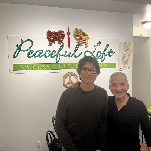 Two men smiling in a vegan restaurant with a sign reading "Peaceful Loft Vegan Restaurant" decorated with festive ornaments and a drawing of a rabbit with the text "Hoppy 2023".