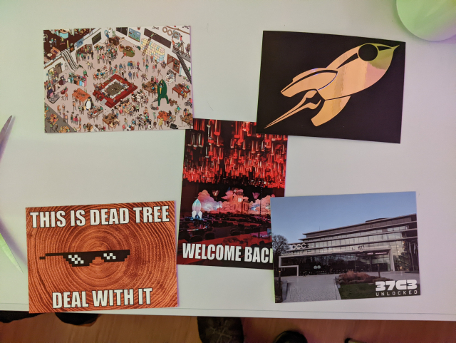 5 postcards on a white table. They are arranged like the dots on a die .

The one in the center shows a view of Saal 3 at 37c3 with the text "Welcome Back" over it.
The top left is a drawing in the style of "Where's Waldo?" depicting Saal 3.
The top right is a fairy dust made from gold foil on a black background.
The bottom left is a varation of the "Deal with it" meme showing a pair of sunglasses on a closeup photo of a tree stump with the rings visible, the text says "This is dead tree, deal with it".
The bottom right shows the CCH with the CCC and the message board on the front displaying the 37c3 graphics.
