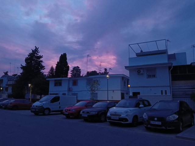 El cielo está con nubes y claros, lo que hace que la luz que atraviesa las nubes se vea rosa y lila. Dado que está poniéndose el sol, el resto de la foto está más oscurecida en un tono azul. Hay tres casas bajas de autoconstrucción con paredes totalmente blancas, tejados planos y ventanas colocadas asimétricamente. Delante de ellas, una fila de coches aparcados frente a la acera, donde ya están las farolas encendidas.