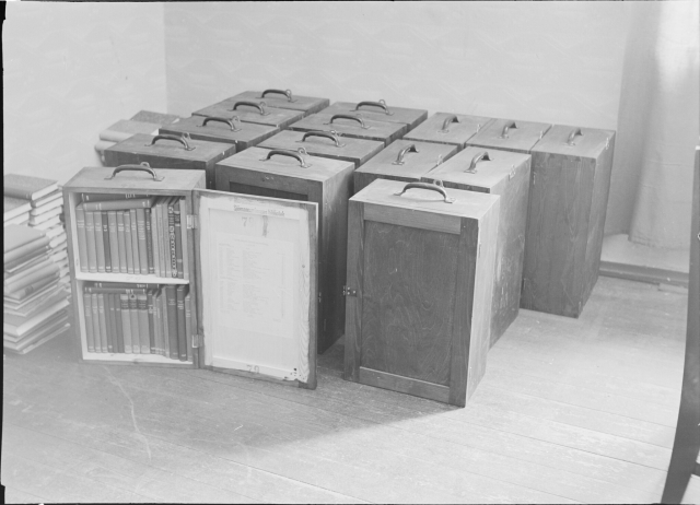 black and white photo of fifteen boxes with handles on the top of them. One in the front is open to reveal two shelves that have about thirty books stacked inside.