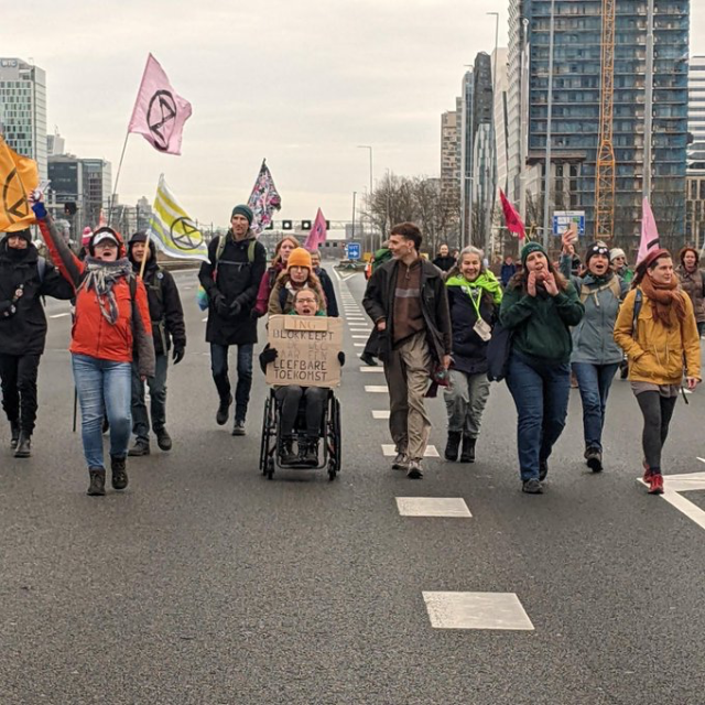 Demonstranten lopen op de A10 met XR-vlaggen. Een van en zit in een rolstoel en houdt een protestbord vast met de tekst 'ING blokkeert de weg naar een leefbare toekomst'. 