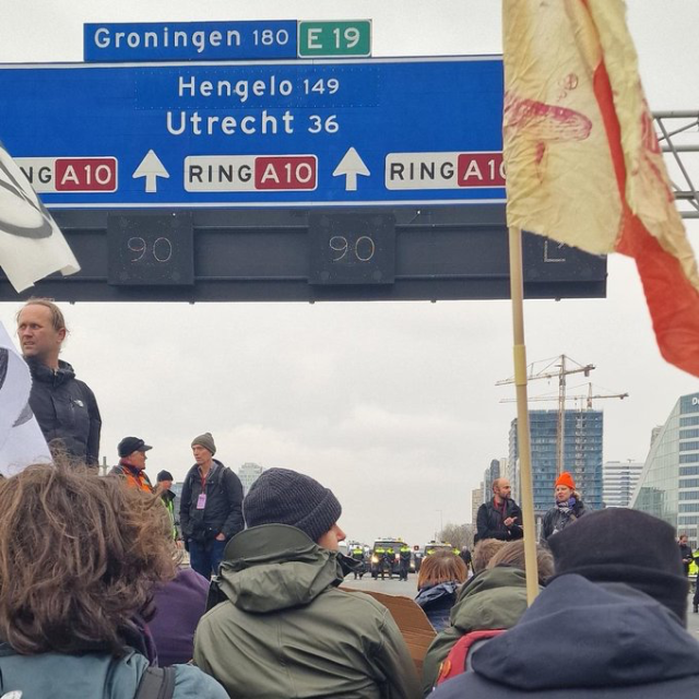 Demonstranten zitten op het asfalt onder blauwe matrixborden. Een aantal van hen houden XR-vlaggen vast. Op de achtergrond politieagenten en politiebussen.