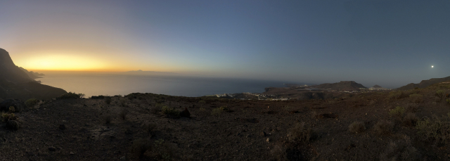 An evening panorama of an evening desert landscape. The sun just set on the left side of the picture, while the moon has just risen on the right. The ground and the sea are list by the rays from both directions, giving them a feeling of unreality. The street lights from the distant towns further away, add a few more dots and lines to the overall picture.