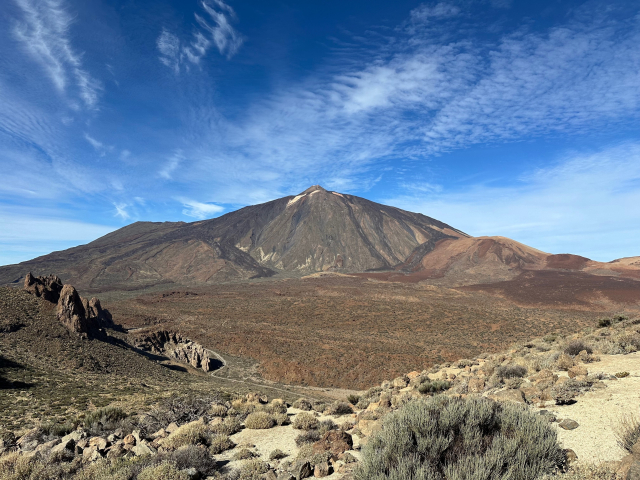 Mountain Teide dominates the landscape of the island of Tenerife. Its perfect conical shape sits in the middle of the picture, under a slight ripple of high altitude clouds on otherwise clear skies. The terrain around and below the mountain resembles a giant bowl covered with the sparse desert vegetation.