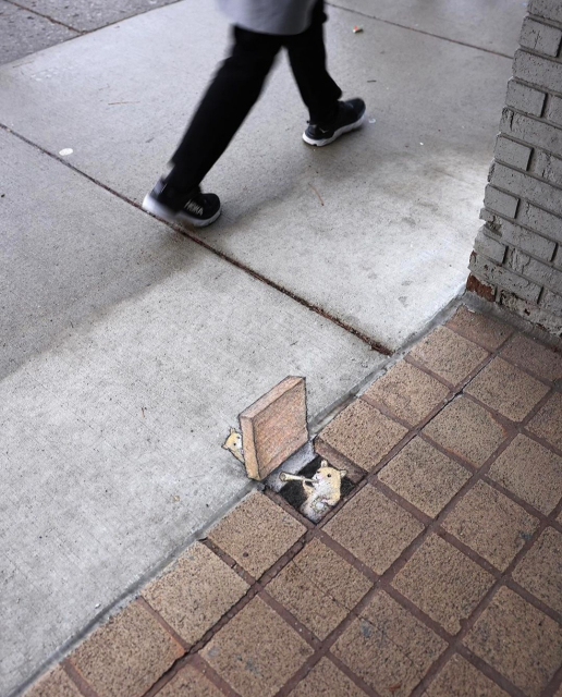 Streetart.  A small hamster has been drawn with chalk on a gray sidewalk. He looks out of a (painted) trapdoor and trumpets into a golden horn. Another hamster stands behind the trapdoor and listens to him. The surroundings consist of small brown tiles, the trapdoor is painted in 3D and in the same way. A passer-by is walking past in the photo.