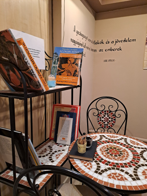 Interior photo of the shop. Round tables and art deco chairs, decorated with colorful mosaics. A bookshelf featuring several books on mythology (and more books under the sugar and coffee bean cups on every table). Quotes painted on the walls.