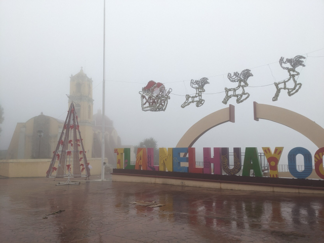 unas letras grandes de colores que dicen "tlalnelhuayocan" al fondo de un patio de concreto rojo. El suelo está todo mojado y hay mucha neblina. Al fondo se aprecia una iglesia y todavía quedan algunos adornos navideños