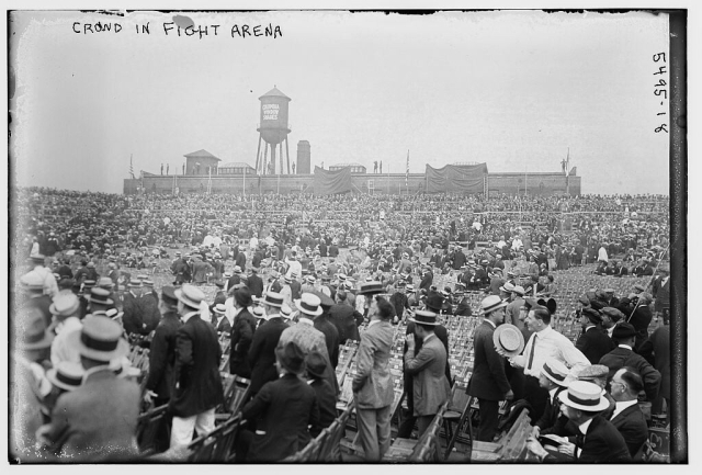 A vast early 20th century crowd standing before a factory whose water-tower and smokestacks rise in the distance.