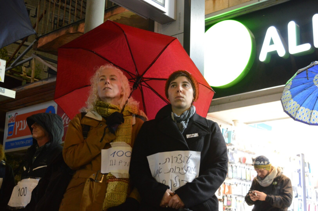 Two people standing under a red umbrella, with signs taped to their clothes. One reads “Stopping. Bringing them back” and the other “100 days”. In the background: commercial street.