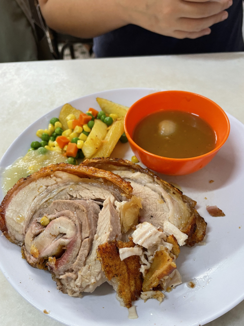 A plate of sliced roasted pork with a side of vegetables and French fries, accompanied by a small bowl of gravy, with a person seated in the background.