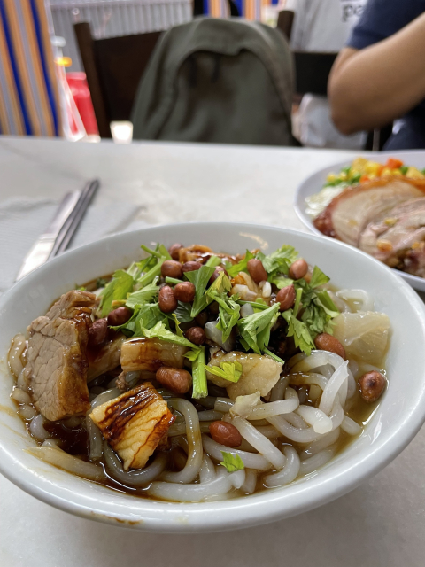 A bowl of noodles with meat, peanuts, and greens, on a table with a fork and spoon at the side, and another dish visible in the background.