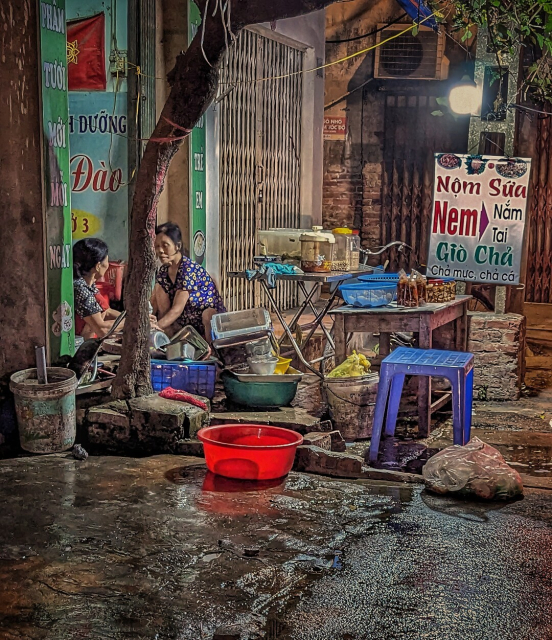 A late evening photo of some sidewalk vendors cleaning up their spot on a small Hanoi street. Two older women in floral print short sleeve shirts and tops kneel behind a tree and wash dishes. All around them are wet sidewalks and wash basins. The table they sold from is still largely intact, though much has been put away for the night in preparation of a complete takedown. Only some pre-packaged goods for a quick takeaway remain displayed for any late arriving customers. Their sign advertises a few dishes common in the nearby coastal province of Nam Định: jellyfish salad, a couple of pork dishes (one meatball-like, another fermented), as well as more familiar spring rolls.
