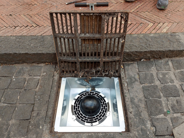 Sewer drain on a street, with the metal lid open. Inside, a metal plate with the black plastic filter in the middle. This setup reduces mosquito populations by 92%.