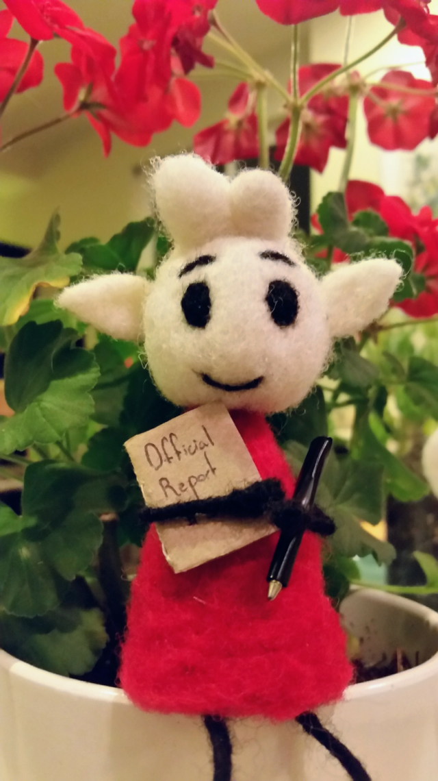 Needle felted Alfur sitting in a geranium pot. he's holding an official report and a pen.