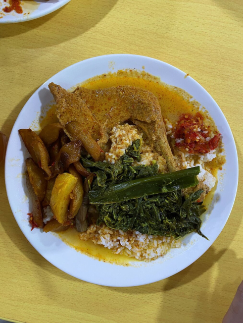 White plate serving white rice, drumstick piece of Ayam Gulai with lots of its gulai poured all over the rice, Sayur Pucuk Paku Masak Lemak, Kentang Cili (Chilli potato wedges), and Sambal.