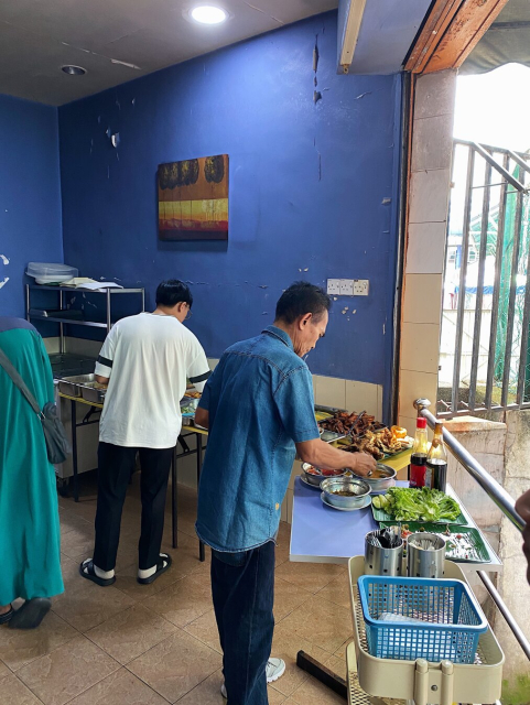 Small corner shop with blue walls, and small tables serving food for people to choose from and take for themselves. Few patrons are queueing while taking their choice of food. Bottles of soy sauce and containers filled with utensils are also placed on the table.