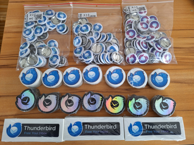 From top to bottom, an assortment of Thunderbird logo pins and stickers. The logo has a stylized blue bird with a white eye curled counter-clockwise around an envelope.