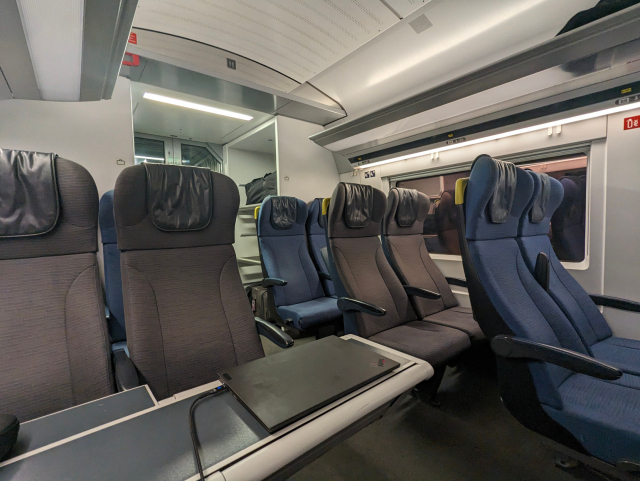 Interior of a train carriage with a variety of seats in a 2-2 configuration. In the foreground, a table with a laptop indicates a workspace. The seats are upholstered in dark and blue fabric with a ribbed texture, and the carriage is well-lit with overhead lighting. Luggage racks above the seats are partially filled. The carriage appears to be nearly empty, providing a quiet travel environment.