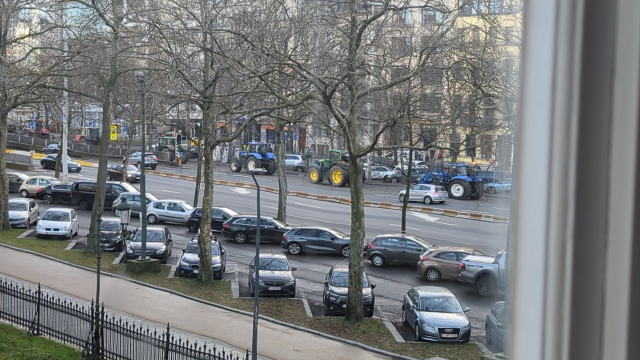 A view from a window overlooking a busy street in Brussels, where a farmers' protest is in progress. Several tractors, easily distinguished by their large size and bright colours, are driving down the street among an assortment of cars.