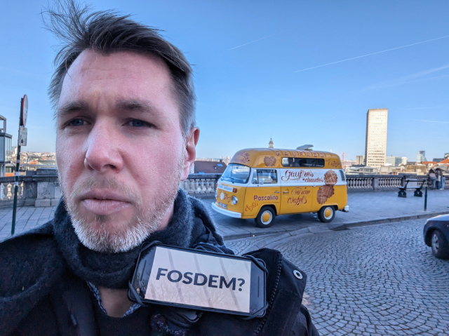 A man stands in the foreground, taking a selfie, with a cityscape behind him under a clear blue sky. He has a questioning expression, and a phone attached to his shoulder with the text "FOSDEM?" displayed on the screen. In the background, there's a vintage yellow van with "Gaufres" written on it, indicating it's a waffle vendor.