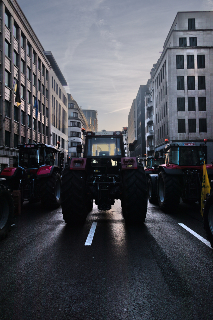 A tractor in the middle of the street in brussels, surrounded by other tractors and with the bright morning sky behind its habitacle