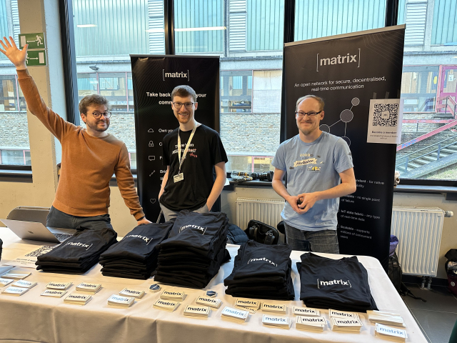 A picture of three persons looking happy at the Matrix booth at FOSDEM, in front of a pile of stickers and T-shirts