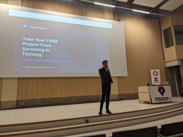 Ryan Sipes from the Thunderbird Project, dressed in all black, takes the FOSDEM Main Stage in front of a slide that says "Take Your FOSS Project From Surviving to Thriving"
