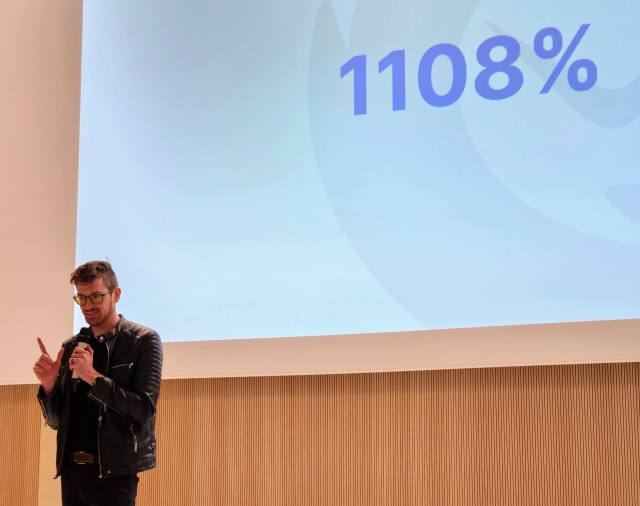 Ryan Sipes from the Thunderbird project speaks during his Main Track FOSDEM talk, pointing upwards while the text "1108%" is visible behind him. Business, business, business, numbers, numbers numbers!