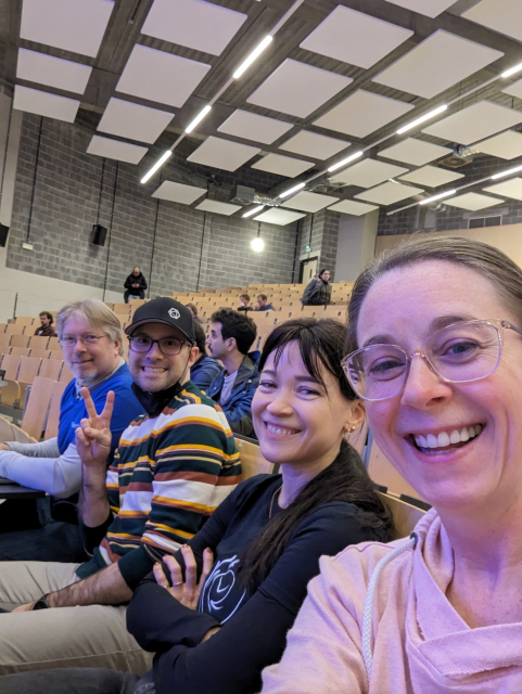 Four members of Team Thunderbird sit in the Main Track room, smiling into the camera, as they wait for Ryan Sipes' talk to start.