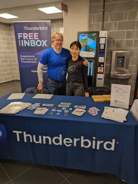 Wayne and Natalie from Team Thunderbird standing behind a fully swagged out table at the FOSDEM stand.
