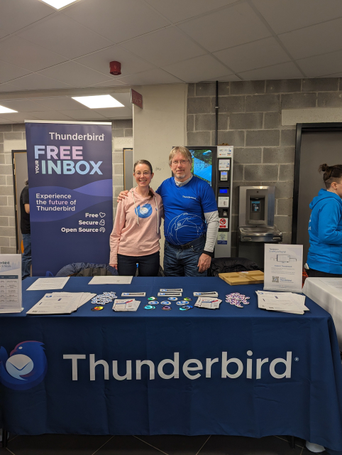 Heather and Wayne standing behind a table laden with Thunderbird stickers and various flyers.