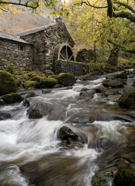 A photo of a stone built watermill. The mill stands next to a fast flowing rocky stream. The motion of the water has been smoothed by a long exposure. Hanging above the stream are golden leaves from and moss covered branches from trees that have grown on either side of the water.
