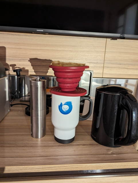 A pourover setup with a hand grinder and kettle, and a red pourover silicone cone over a white travel mug with a Thunderbird logo.