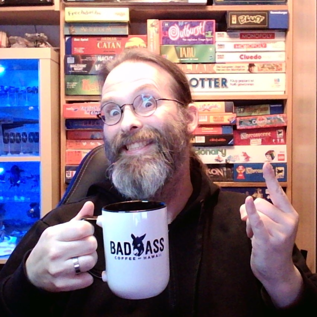 Bearded old man with a mug saying "Bad Ass" on it making the metal horns.