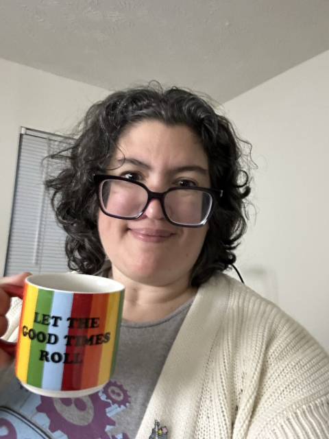 A smiling person wearing glasses and a cream-colored cardigan holds a colorful mug with the phrase "LET THE GOOD TIMES ROLL" printed on it.
