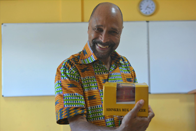 Photo of a black man standing in a classroom, smiling, and holding a box containing a mug