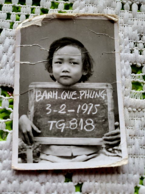 Me, in refugee mugshot at age 3. Holding chalkboard with my name,  birth date & refugee camp number.