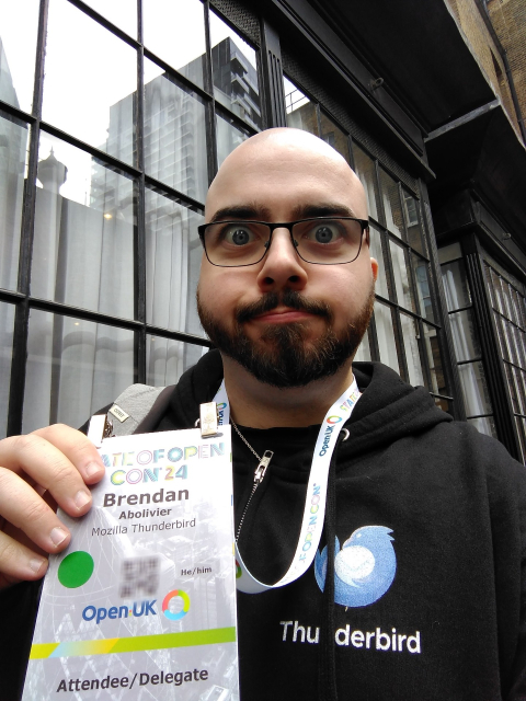A selfie I took while wearing a hoodie featuring the Thunderbird logo, and holding a badge for the State of Open 2024 conference with my name and "Mozilla Thunderbird" under it.