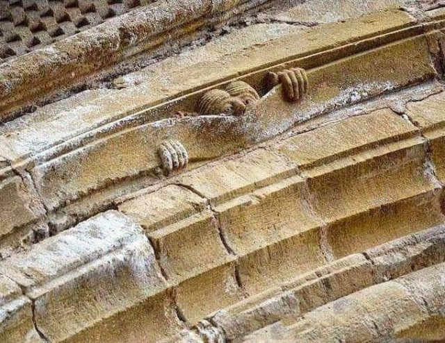 The layered arch of a building includes a sculpture of a man trapped in the stonework, just the top of their head and one hand poking out. Possibly the earliest recorded Kilroy.