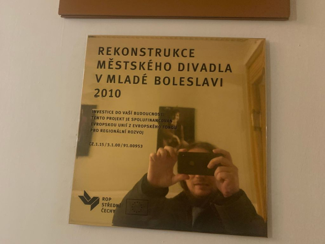 Nemlady jiz pan neuspesne imituje influencery fotici se v zrcadle. Na zlate plakete je napsano: "Rekonstrukce Mestskeho divadla v Mlade Boleslavi 2010."