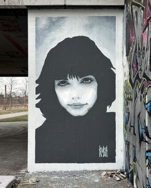 Streetartwall. A simple but beautiful black and white portrait of a young woman was sprayed on a wall next to a road bridge. She has dark short hair, dark eyes, full lips and a small nose. She is wearing a black top. Her gaze is friendly and directed at the viewer. Next to the mural is part of a wall with graffiti and a view of a path under the bridge to buildings in the distance. 