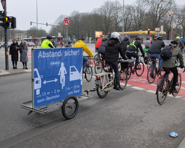 Ride of Silence mit mehreren Radfahrern; Anhänger mit Schild das sichere Überholabstände zeigt