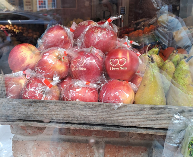 A pile of red apples in a produce store window. Each apple is wrapped in plastic, and into their skin two hearts are carved, with the words "I love you".
