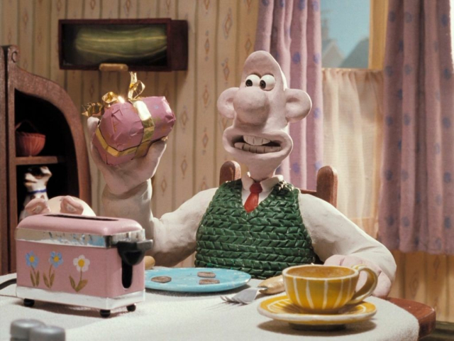 A man made out of clay sits at a table. His head is bald and his ears stick out. He's wearing a green sweater vest and a red tie. In his hand is a pink present wrapped with a shiny gold bow.