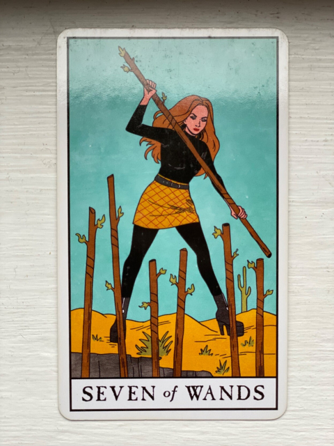 Seven of Wands from the Modern Witch Tarot: a white woman with long hair and neat makeup stands at the top of a cliff, wooden staff held in both hands to knock back six more staffs that rise up from below to challenge her